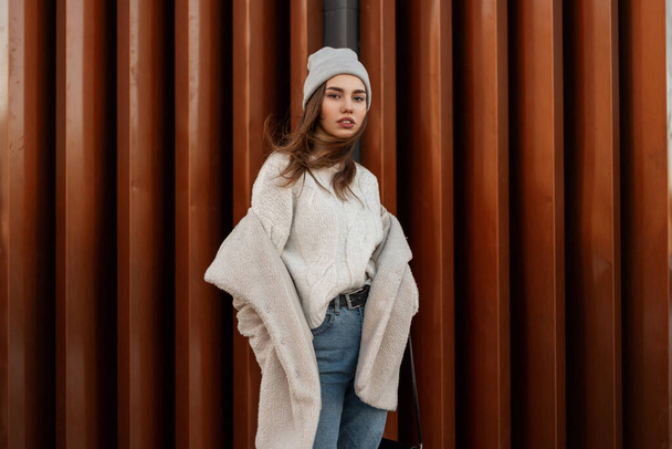 Pretty stylish young woman model in a fashionable knitted sweater in a knitted hat in an artificial fur coat in jeans stands near a vintage building outdoors. Cute trendy modern girl in youth clothes. - Photo, Image