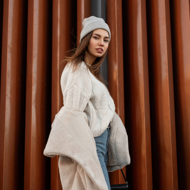 Beautiful pretty young woman in a stylish knitted sweater in a fashionable warm hat with a youth coat of their faux fur posing outdoors in the city near a modern wall. Attractive girl. Spring style. - Zdjęcie, obraz