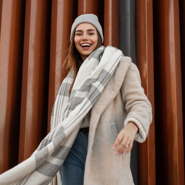 Cheerful girl with funny smile in trendy clothes outdoors. Positive young woman in coat in a knitted hat in a woolen vintage warm scarf stands and beautiful smiles near a modern wall in the street. - Photo, image