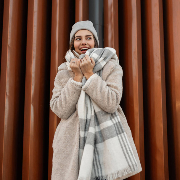 Modemodel van een mooie vrolijke jonge vrouw met een leuke glimlach in modieuze nepbontjas in een gebreide stijlvolle muts met een wollen geruite sjaal poserend bij een metalen muur in de stad. Vrolijk meisje. - Foto, afbeelding