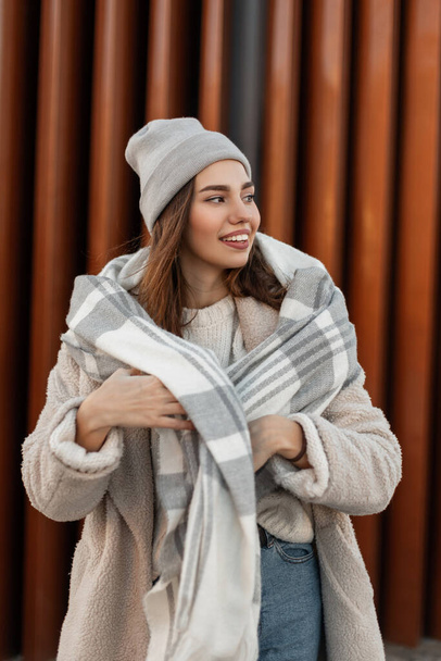 ウールの暖かいスカーフを持つ魅力的なフェイクファーのコートでスタイリッシュなニット帽の楽しい若い女性モデルが立っており、通りの近代的な建物の近くの側面に見えます。かわいいです面白い女の子上の通り - 写真・画像