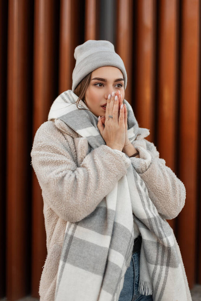 Europese mooie jonge vrouw met een stijlvolle hoed in een eco jas met een wollen warme vintage sjaal bedekt haar gezicht met haar handen in de buurt van een moderne muur. Urban aantrekkelijk meisje model poseren buiten in de stad. - Foto, afbeelding
