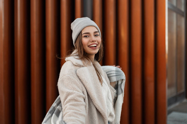 Mujer joven glamorosa bastante feliz en un sombrero vintage de punto en un abrigo de piel sintética de moda con bufanda de lana de moda se encuentra cerca de un edificio moderno en la calle. Chica alegre modelo al aire libre en la ciudad. - Foto, imagen