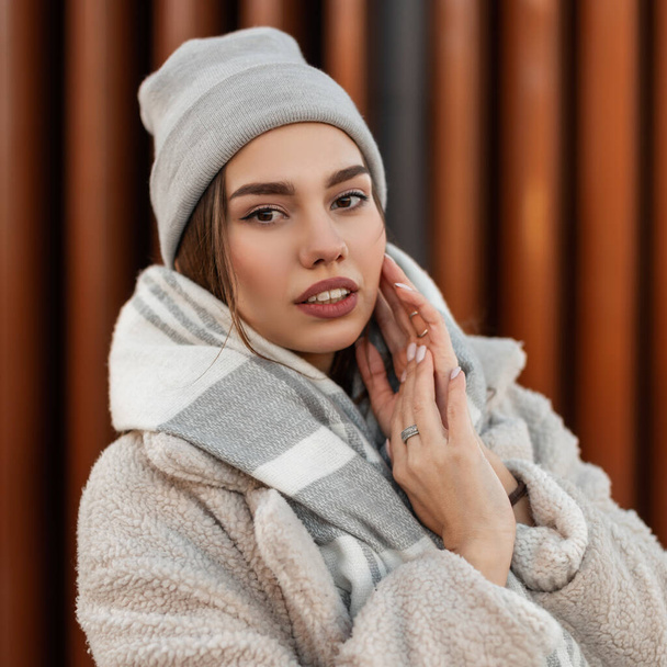 Retrato de una hermosa mujer joven con labios sexy en un sombrero de punto de moda en un abrigo con una bufanda a cuadros cálida y elegante de lana al aire libre. Linda chica en ropa de moda en la calle cerca de una pared moderna - Foto, Imagen