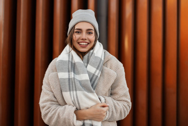 Portrait of a cute young woman with a positive smile in a fashionable knitted hat in an elegant faux fur coat with a vintage woolen plaid scarf near a modern wall in the city. Cheerful girl outdoors. - Photo, Image
