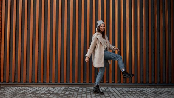 European joyful young woman with a beautiful smile steps near a metal vintage wall in the city. Urban funny girl model in trendy clothes walks along the street. Youth women's stylish spring clothing. - Photo, image