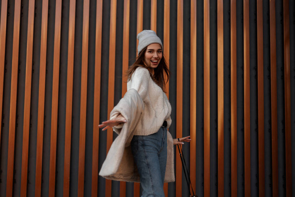 Привлекательная позитивная молодая женщина с красивой улыбкой в модной шляпе в трикотажном свитере в джинсах позирует на фоне современной городской стены. Милая весёлая девушка-модель обманывает. - Фото, изображение