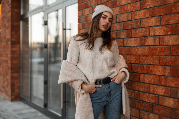 Young stylish woman in youth warm fashionable outerwear is resting near a vintage brick building outdoors on a spring day. Attractive girl fashion model in trendy clothes enjoys relaxes in the street. - Photo, Image