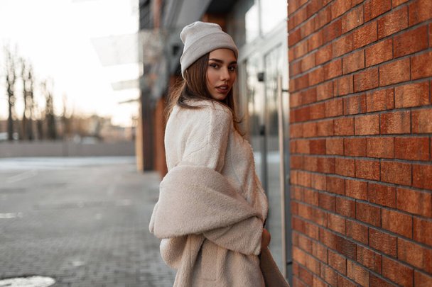 Beautiful young woman in a fashionable gray hat in a stylish knitted sweater in a youth spring eco fur coat posing near a vintage brick building on the street. Attractive girl model walks on the city. - Foto, immagini