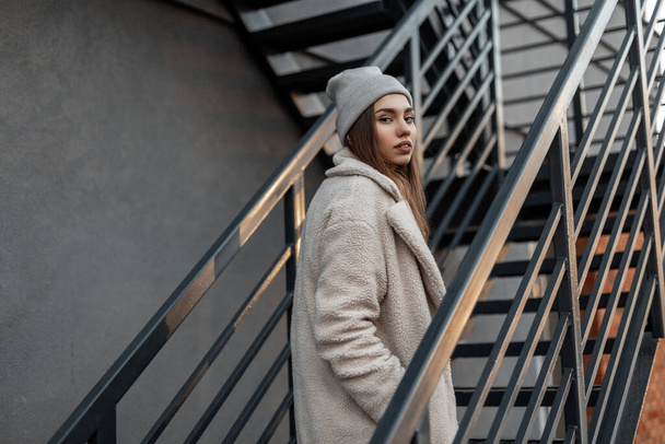 Young trendy European woman in a fashionable knitted warm hat in a stylish fur coat made of faux fur is standing in the city on a metal vintage staircase. Modern girl model outdoors. - Fotó, kép