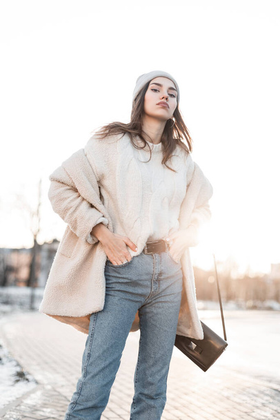 Beautiful young woman in trendy warm winter clothes is enjoys the bright sunset on a street in the city. Cute girl fashion model on a walk in a winter day. Stylish clothes for women. New collection - Photo, Image