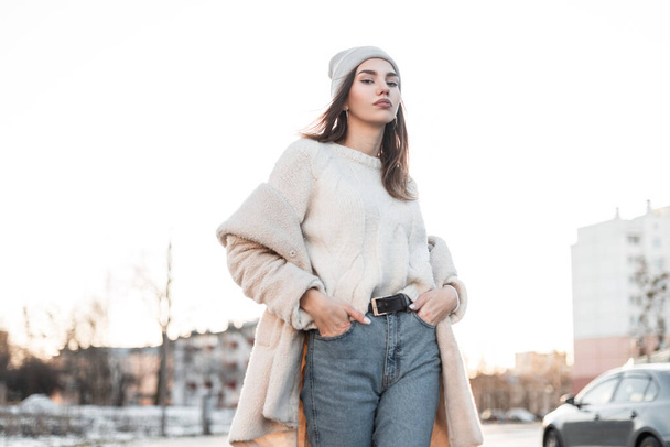 Mujer joven europea en un sombrero de punto en un suéter caliente en un abrigo de piel sintética de moda posando al aire libre en un día de invierno soleado brillante. Modelo de chica atractiva en ropa de moda al aire libre. Buen día soleado. - Foto, Imagen