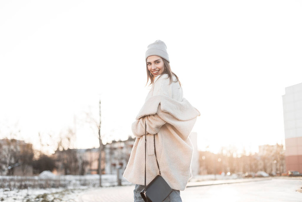 Happy beautiful young woman model with funny smile in a fashionable warm vintage knitted hat in an eco fur coat poses outdoors in the city. Cheerful girl in youth winter clothes walks on the street. - Photo, Image