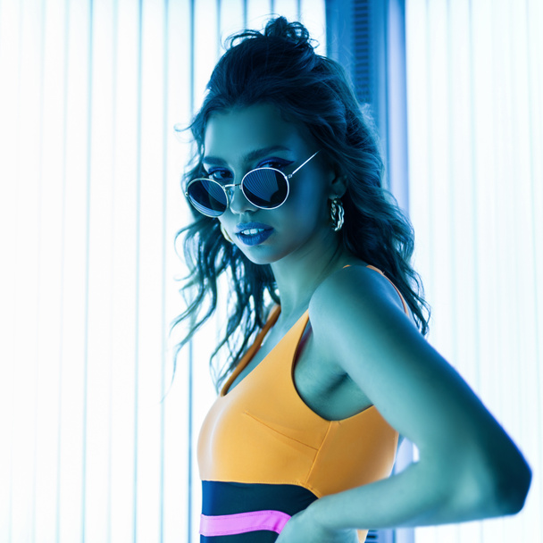 Stylish sexy young woman with curly hair in trendy sunglasses in a colored stylish swimsuit sunbathes in a solarium. Attractive hipster girl is standing in a modern tanning bed. - Fotografie, Obrázek