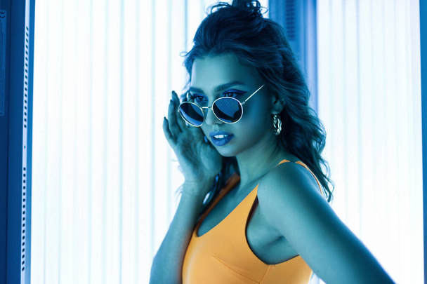 Young glamorous hipster woman in fashion sunglasses with a trendy hairstyle in a yellow stylish swimsuit is standing in the solarium and enjoys the ultraviolet.Sexy girl sunbathes in a modern solarium - Photo, Image