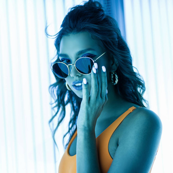 Portrait of a young woman in curly hair in sunglasses with a stylish manicure in a trendy yellow swimsuit in a modern solarium. Sexy hipster girl adjusts glasses and enjoys the ultraviolet light. - Photo, Image