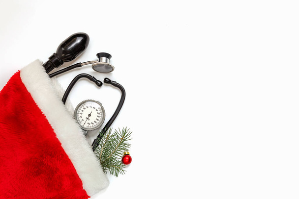 Biglietto di Natale medico con uno stetoscopio e un ramo di abete rosso in un cappello di Babbo Natale su sfondo bianco. La medicina di anno nuovo. Ricevuto. Posa piatta. - Foto, immagini