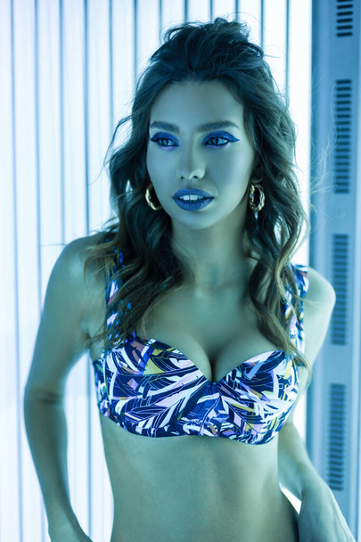 Sexy sensuele jonge vrouw met krullen met gezondheid gebruinde huid met blauwe lippen in een stijlvol zwempak staat in een solarium met moderne ultraviolette lampen. Elegant meisje en neon licht in een zonnebank. - Foto, afbeelding