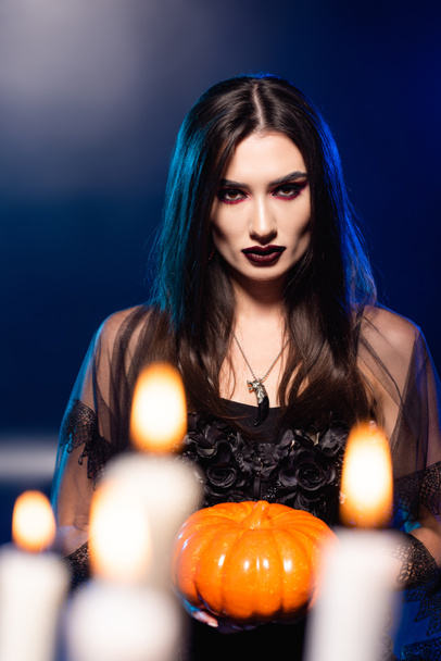 selective focus of young woman with black makeup holding pumpkin near burning candles on blue, halloween concept - Φωτογραφία, εικόνα