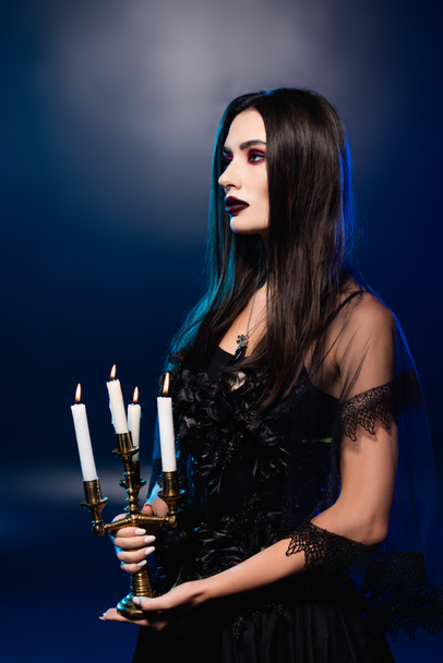 pale woman with black makeup holding candlestick with burning candles on blue, halloween concept - Photo, Image