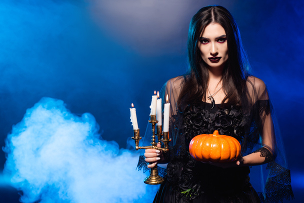 young pale woman with scary makeup holding pumpkin and burning candles on blue with smoke, halloween concept - Photo, Image