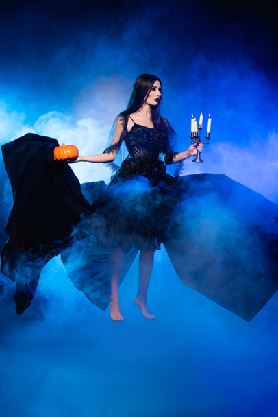 jong blootsvoets vrouw in zwart jurk houden pompoen en branden kaarsen op blauw met rook - Foto, afbeelding