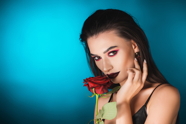młoda kobieta z czarnym makijażem w pobliżu czerwonej róży na niebiesko - Zdjęcie, obraz