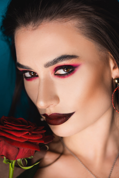 close up of young woman with dark makeup near red rose on blue - Φωτογραφία, εικόνα