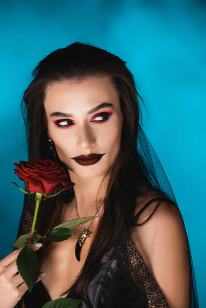 tajemná mladá žena s černým make-up v závoji dívá pryč blízko růže na modré - Fotografie, Obrázek