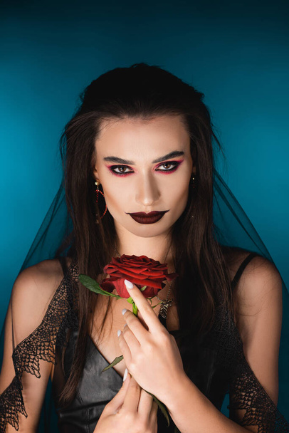 mysterious young woman with black veil and makeup holding rose on blue - Foto, Imagem