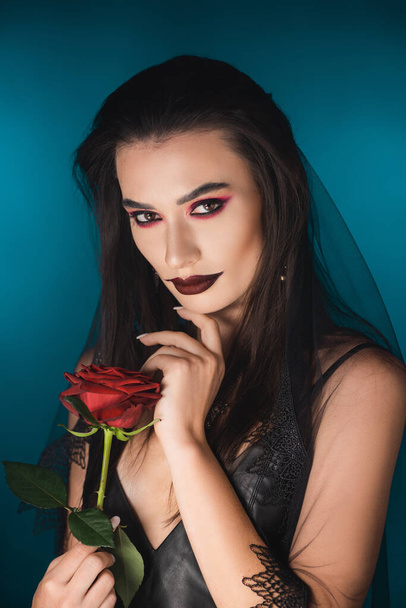 mysterious young woman with black veil and makeup holding red rose on blue - Φωτογραφία, εικόνα