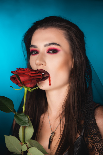 young and creepy woman with blood on face near red rose on blue - Foto, immagini