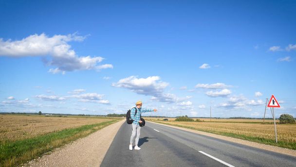 A young man is hitchhiking around the country. The man is trying to catch a passing car for traveling. The man with the backpack went hitchhiking to south. - Photo, image