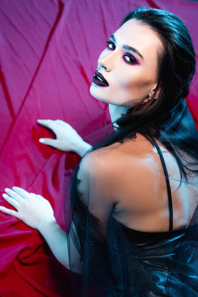 pale and young woman with black makeup looking at camera on red - Foto, Bild