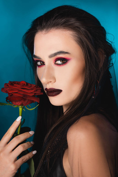 young brunette woman with dark makeup looking at camera near red rose on blue - Foto, Imagem