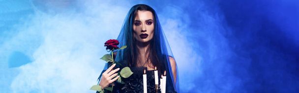 panoramic crop of woman in black veil holding rose and burning candles on blue with smoke, halloween concept - Fotografie, Obrázek