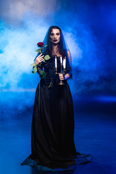 bride in black dress and veil holding  rose and burning candles on blue with smoke, halloween concept - Φωτογραφία, εικόνα