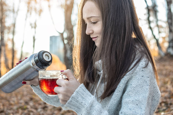 夕日に照らされた秋の黄色い公園で、若い可愛い女性が魔法瓶から熱いお茶を注いでいます。秋の寒い季節に居心地の良いライフスタイルの概念 - 写真・画像