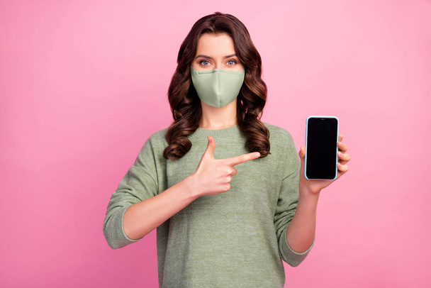 Portrait of positive cheerful girl promoter point index finger smartphone wear medical mask jumper isolated over pastel color background - Photo, Image