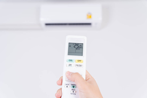 人間の手は、部屋の中のエアコンのオンまたは調整温度のためのエアコンの白いリモコンを使用しています. - 写真・画像