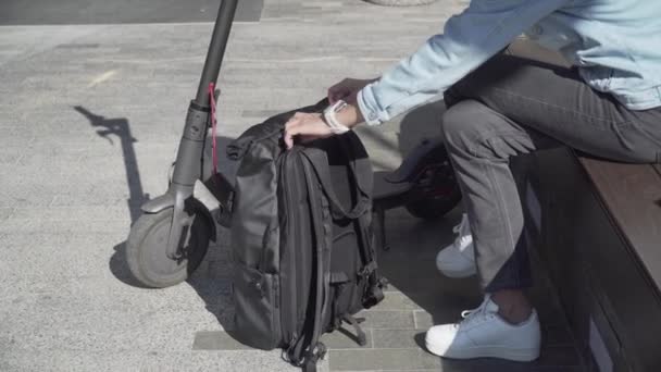 Kafkaslı yetişkin bir kadın şehirde bankta otururken elektrikli scooter kullandıktan sonra sırt çantasından güç bankasını alarak şarj ediyor. Dış güç bankasının pili şarj ediliyor - Video, Çekim