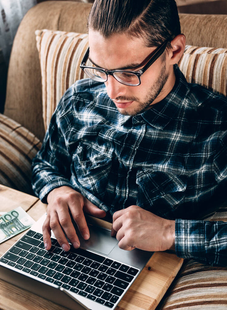 Un joven guapo con una camisa azul y gafas está trabajando en una computadora plateada y el papel moneda Euro está al lado. Trabajo remoto durante el aislamiento y la cuarentena. - Foto, imagen