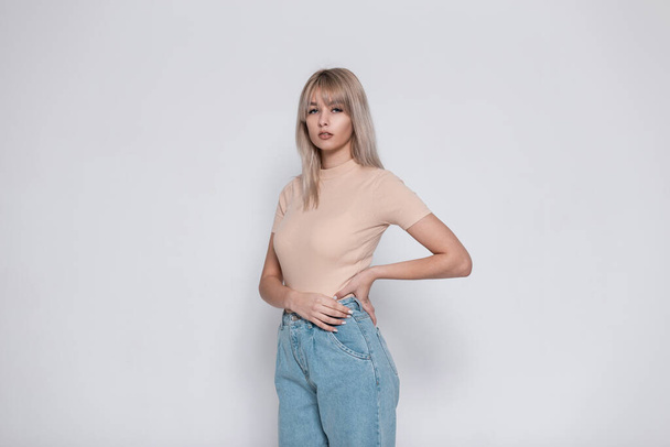 Elegante modello di una giovane donna moderna con i capelli biondi in una t-shirt alla moda beige in jeans eleganti blu in posa vicino a un muro bianco all'interno.Attraente ragazza in abiti alla moda è in stanza.Lady - Foto, immagini