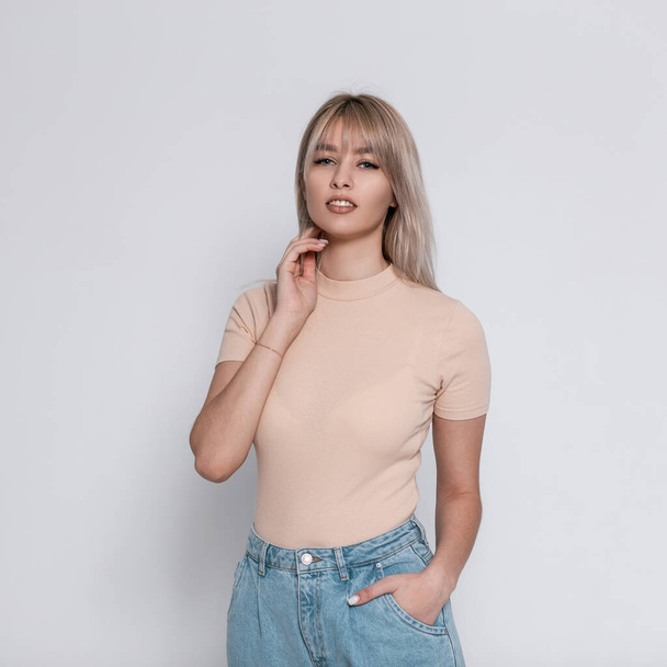 Glamorous fashion model of a young blonde woman in stylish blue jeans in a trendy shirt with a hairstyle stands near a white wall in the room. Cute beautiful elegant girl in youth fashionable wear.  - Фото, зображення