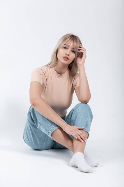 Stylish young woman blonde with trendy hairstyle in fashionable youth casual wear sits near a white wall in the studio. European trendy girl model posing indoors. Spring fashion. - Photo, Image