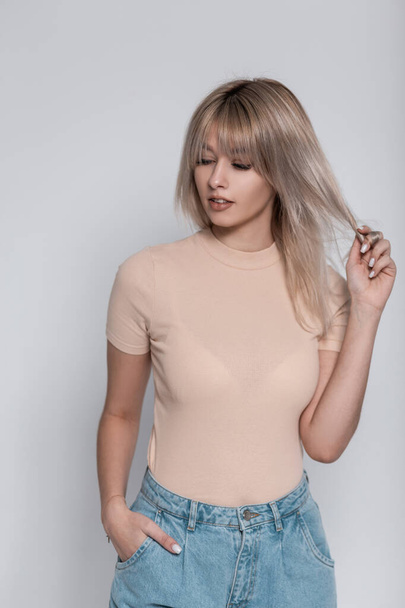 Stylish pretty young woman blonde in fashionable clothes stands and straightens hair near a white wall in the studio. Girl fashion model in a vintage T-shirt in blue trendy jeans posing in the room. - Foto, afbeelding