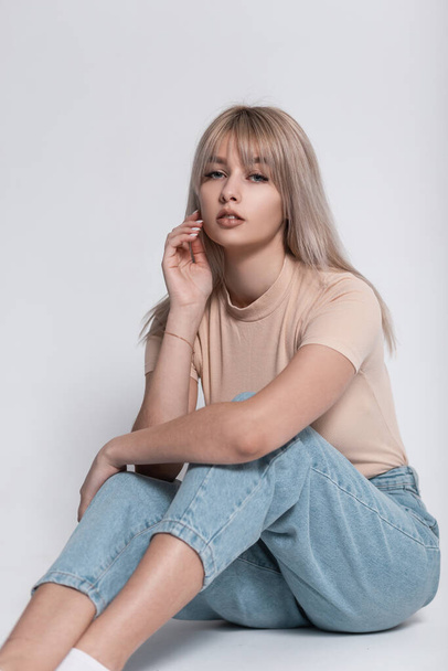 Portrait of a fashionable young blonde woman with a stylish hairstyle in a vintage T-shirt in blue trendy jeans on a white background in the room. Elegant girl fashion model sits on the floor indoors. - Foto, Bild
