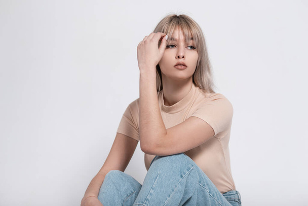 Fashionable young model of a glamorous blonde woman in a stylish t-shirt in trendy jeans is resting sitting near a vintage white wall in the room. Cute european girl model relaxes indoors. Fashion. - Foto, Imagem
