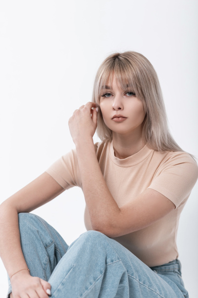 Pretty modern young blond woman in a fashionable beige t-shirt with a stylish hairstyle poses near a white wall in the room. Attractive trendy girl fashion model rests indoors. Elegant cute lady. - Photo, Image