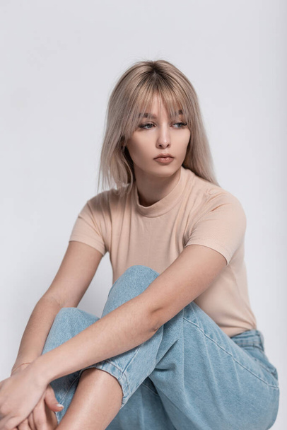 Pretty young attractive woman model in fashionable clothes is sitting near a white vintage wall. European glamorous blonde girl in stylish casual wear is resting in the room. Spring trendy look. - 写真・画像